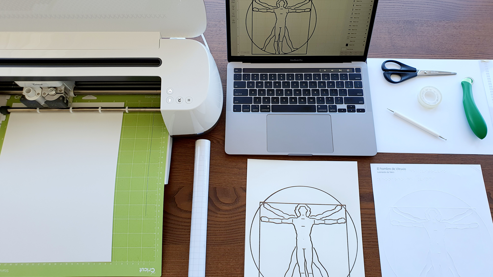 A flat-lay photograph of a desk with a white Cricut Maker cutting machine on the left, a laptop displaying a Vitruvian Man outline, scissors, tape, a green scoring stylus, and two sheets of paper printed with a tactile Vitruvian Man line drawing.